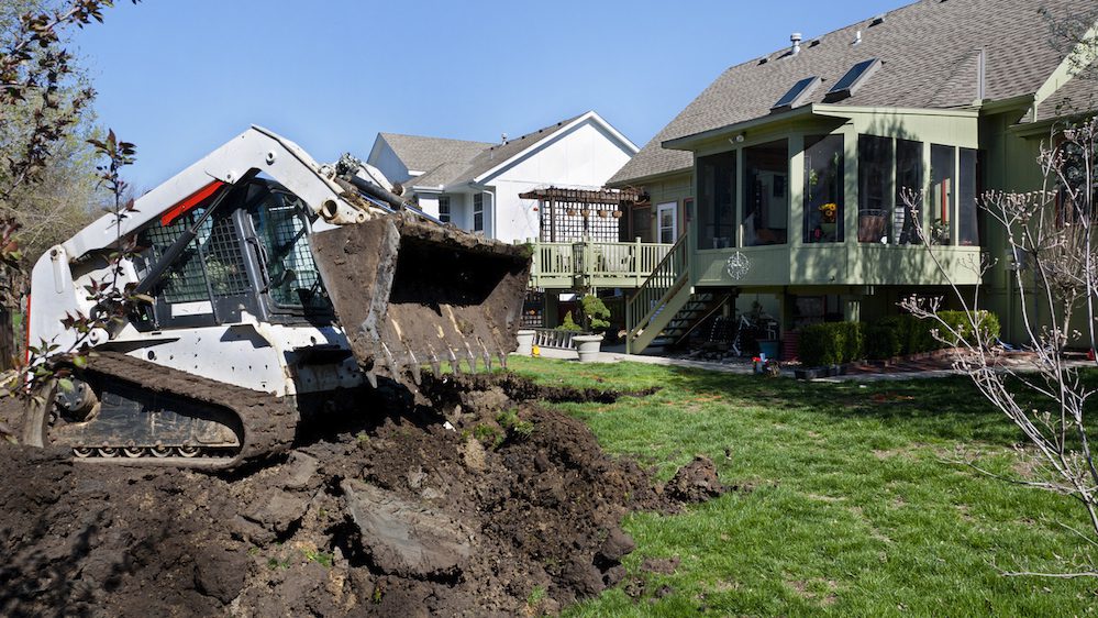 A small digger drops dirt removed from an excavation in back of a suburban house.